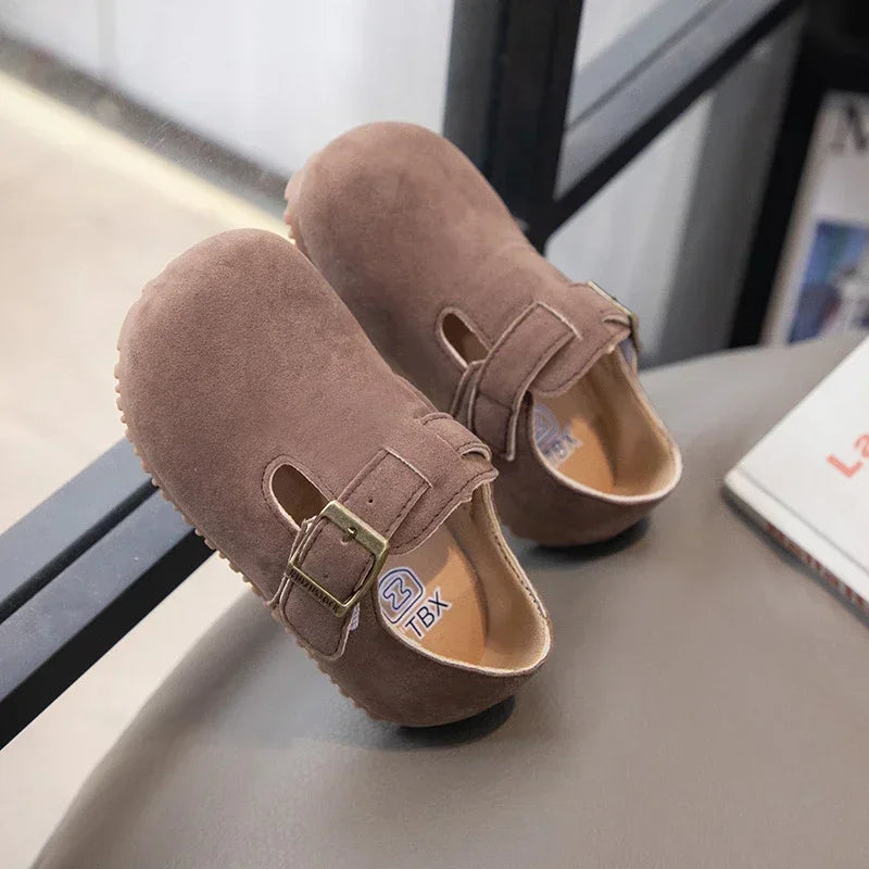 A pair of brown suede kids' moccasin shoes with an easy hook-and-loop closure resting on a gray surface.