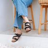 A person wearing black cork footbed slide sandals with arch support, sitting on a wooden stool.