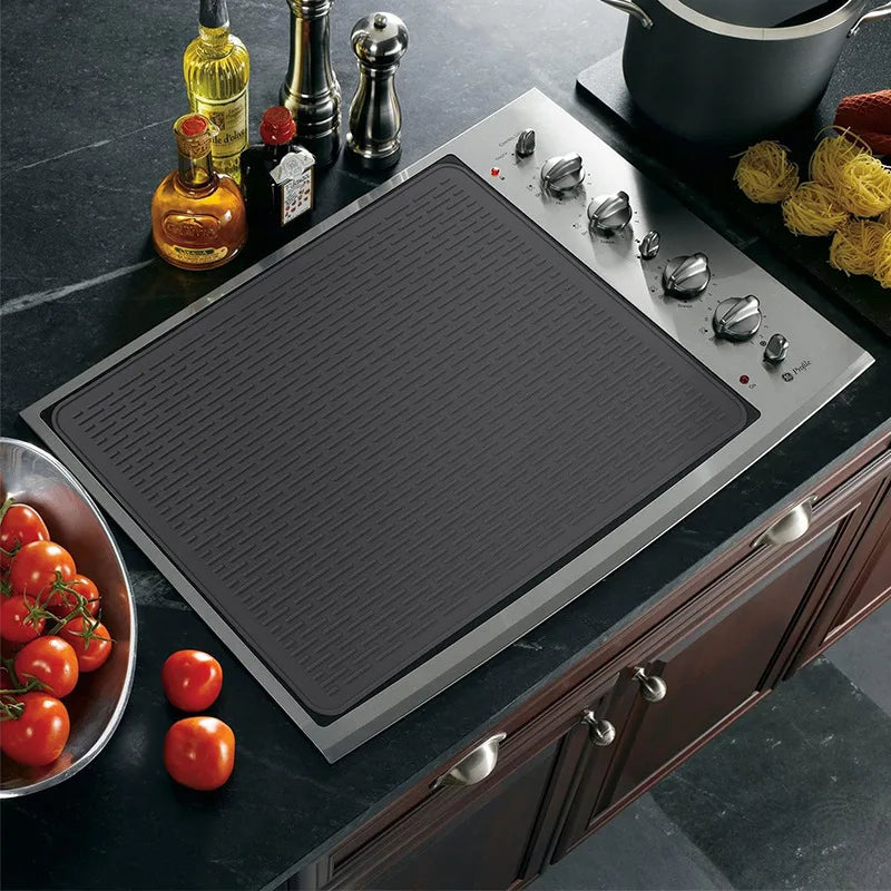 A silicone stove protector mat is placed on a black induction cooktop, surrounded by cooking ingredients and oils.