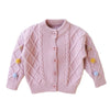 A pink knitted cardigan sweater for girls featuring a diamond pattern and pom-pom trim.