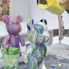 A colorful, painted bear figurine is being dipped in yellow paint while other paint pots and a green container are visible.