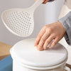 A hand is holding a white long-handled kitchen strainer spoon over a white ceramic pot.