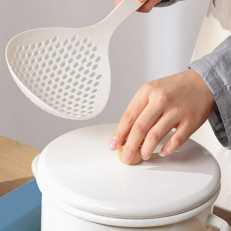 A hand is holding a white long-handled kitchen strainer spoon over a white ceramic pot.