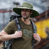 A man wearing a olive green wide brim mesh bucket hat and a t-shirt, carrying a backpack outdoors.