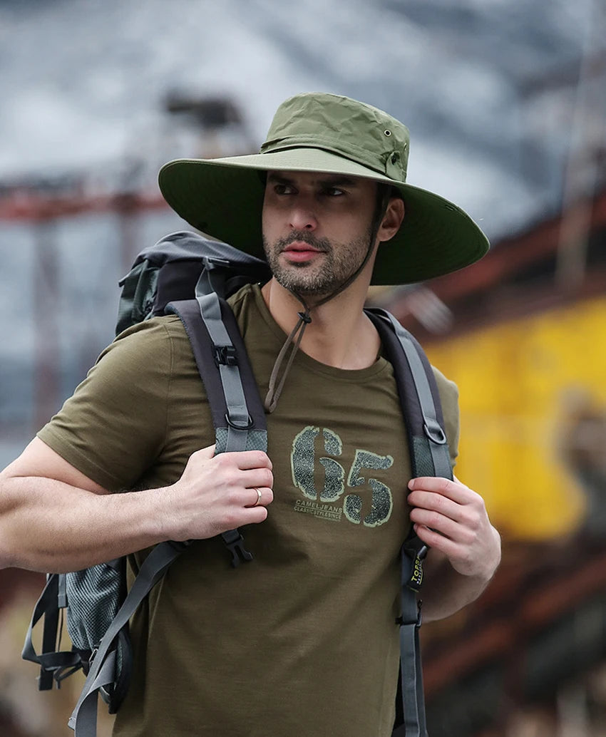 A man wearing a olive green wide brim mesh bucket hat and a t-shirt, carrying a backpack outdoors.