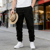A man wearing black cargo pants, a white shirt, and white sneakers, holding a straw hat on a city street.