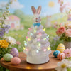Figurine de lapin de Pâques en céramique blanche, décorée de lumières colorées, posée sur une surface en bois entourée de fle