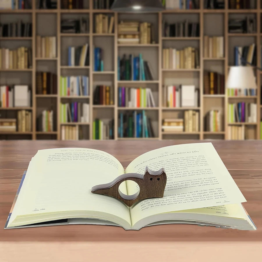 Un libro abierto con un marcapáginas de madera con forma de gato y perro sobre una mesa de madera, con estanterías al fondo.