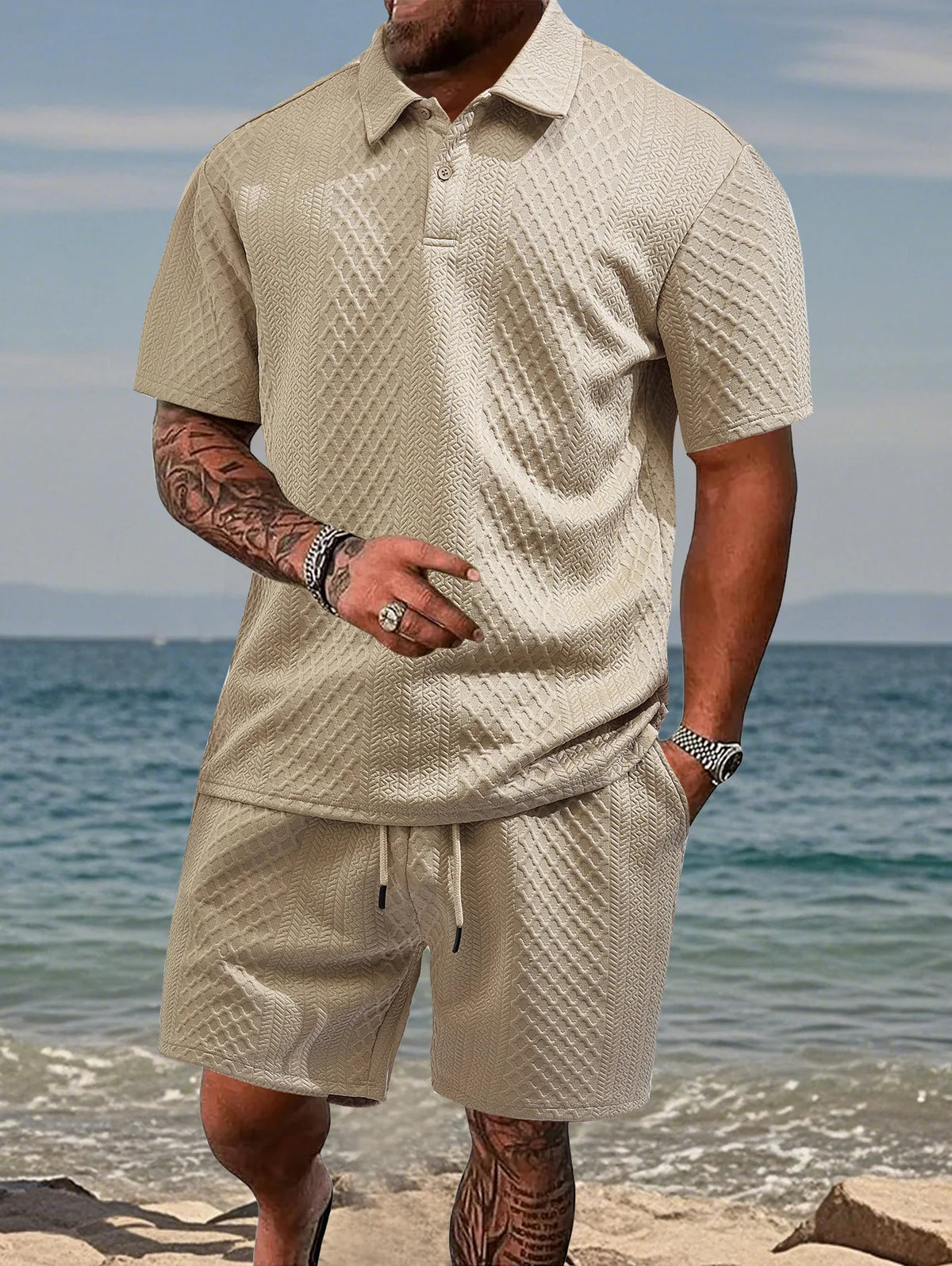 A man wearing a beige jacquard short-sleeve shirt and drawstring shorts set standing near the water.