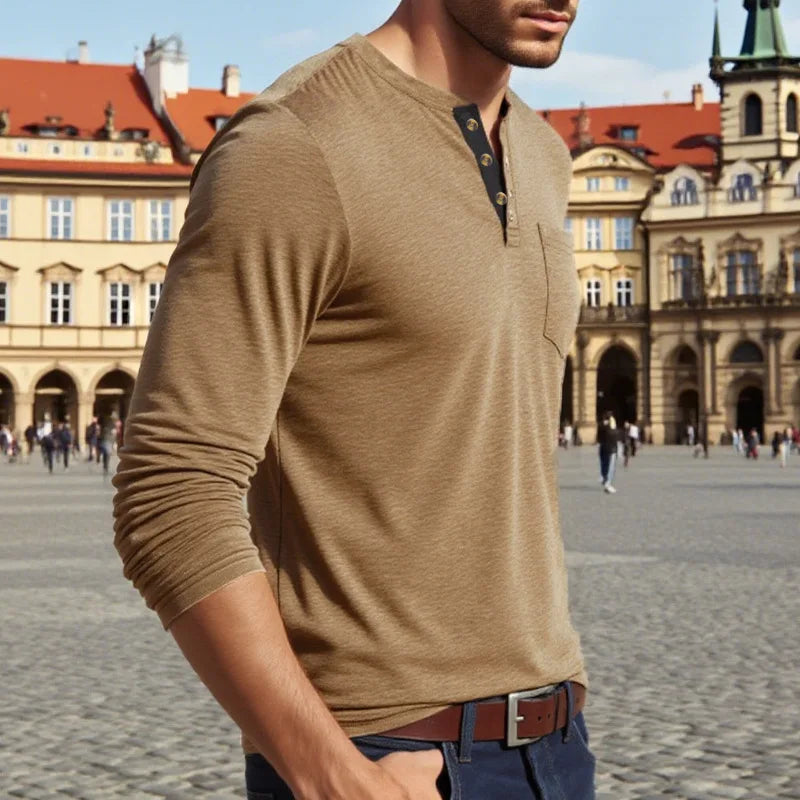 A man wearing a tan long-sleeve shirt with a collar and chest pocket standing in a historic European square.