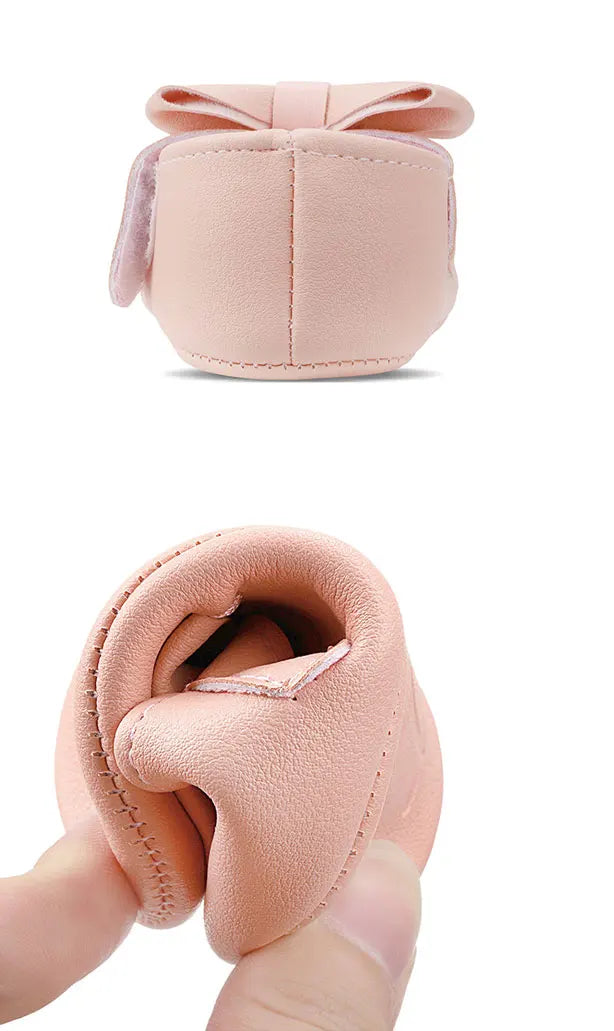 A light pink baby shoe with a bow knot design and anti-slip sole is shown being held by a hand.