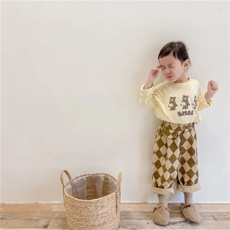 A young child wearing beige patterned harem pants and a yellow top stands against a white wall.