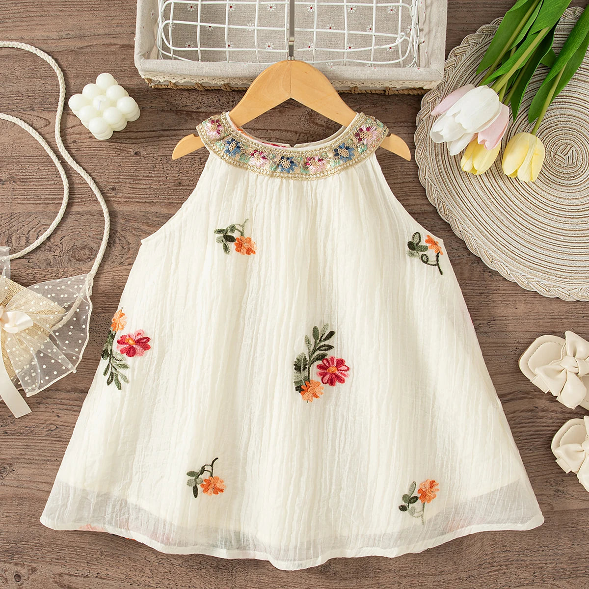 A white sleeveless dress for baby girls with floral embroidery and a delicate lace trim, displayed on a wooden surface.