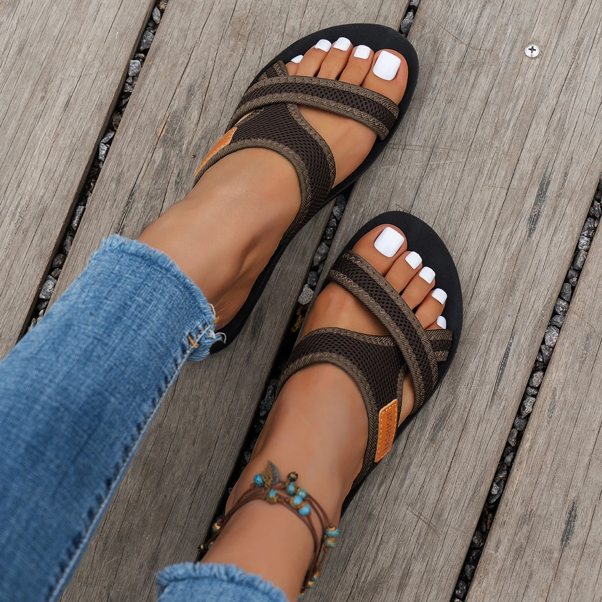 A close-up of women's brown and black breathable mesh flat sandals worn on a foot against a wooden surface.