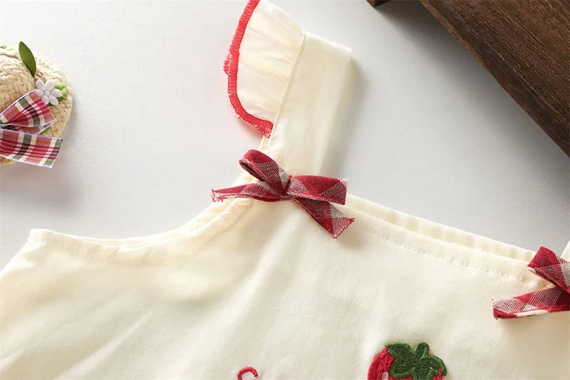 A cream-colored kids summer pinafore dress with red trim and strawberry and bunny print details.