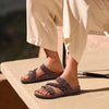 A pair of taupe cork footbed slide sandals with woven straps resting on a light-colored surface.