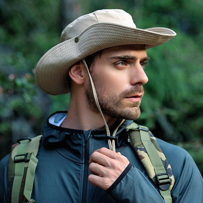 A man wearing a beige waterproof leather rope bucket hat and a dark jacket with a camouflage backpack in an outdoor setting.