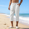 A man wearing white linen drawstring capri pants standing on a beach with ocean waves in the background.