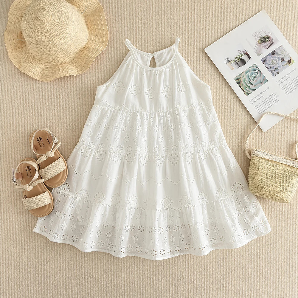 A white cotton dress with eyelet details and a gathered bodice, laid out on a beige fabric next to a straw hat and some shoes