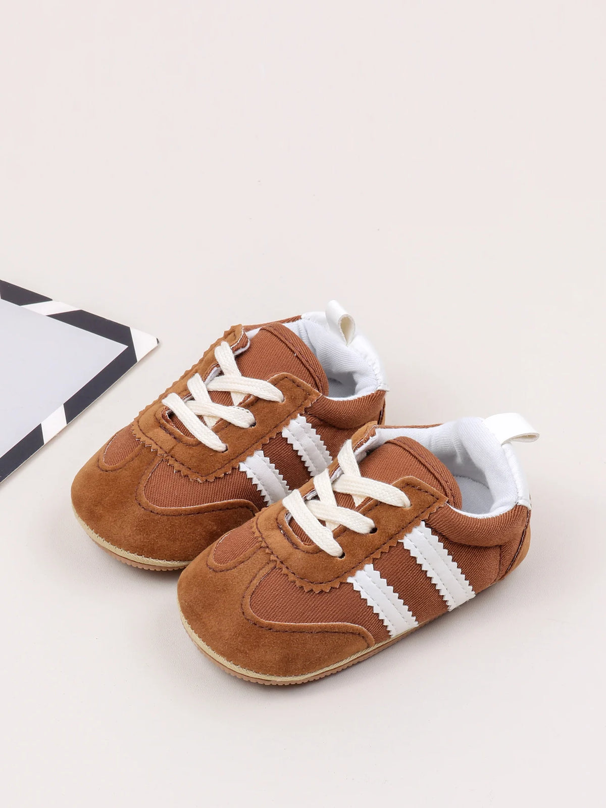 A pair of brown suede baby first walker shoes with white stripes and white soles resting on a light background.