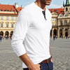 A man wearing a white long-sleeve shirt with a chest pocket and dark blue jeans, standing in front of historical architecture