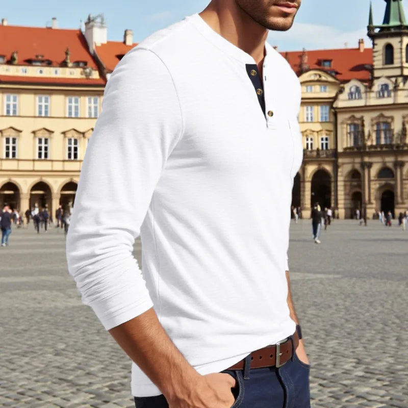 A man wearing a white long-sleeve shirt with a chest pocket and dark blue jeans, standing in front of historical architecture