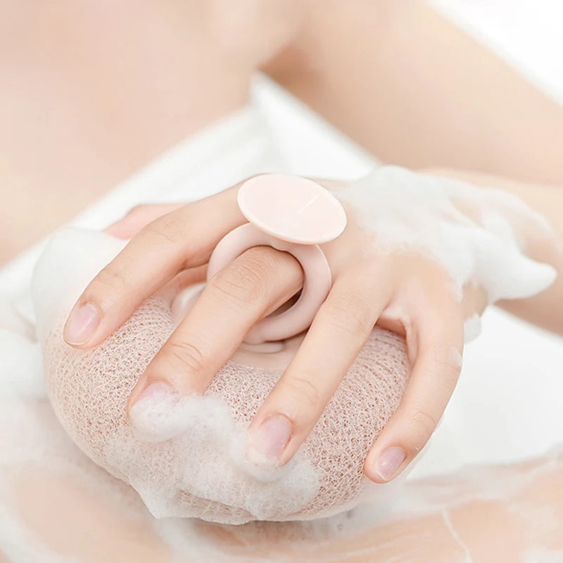 A hand is holding a pink bath sponge with a suction cup and white foam on a light-colored surface.