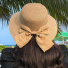A woman wearing an elegant beige straw sun hat with a large bow on her dark hair against a tropical background.