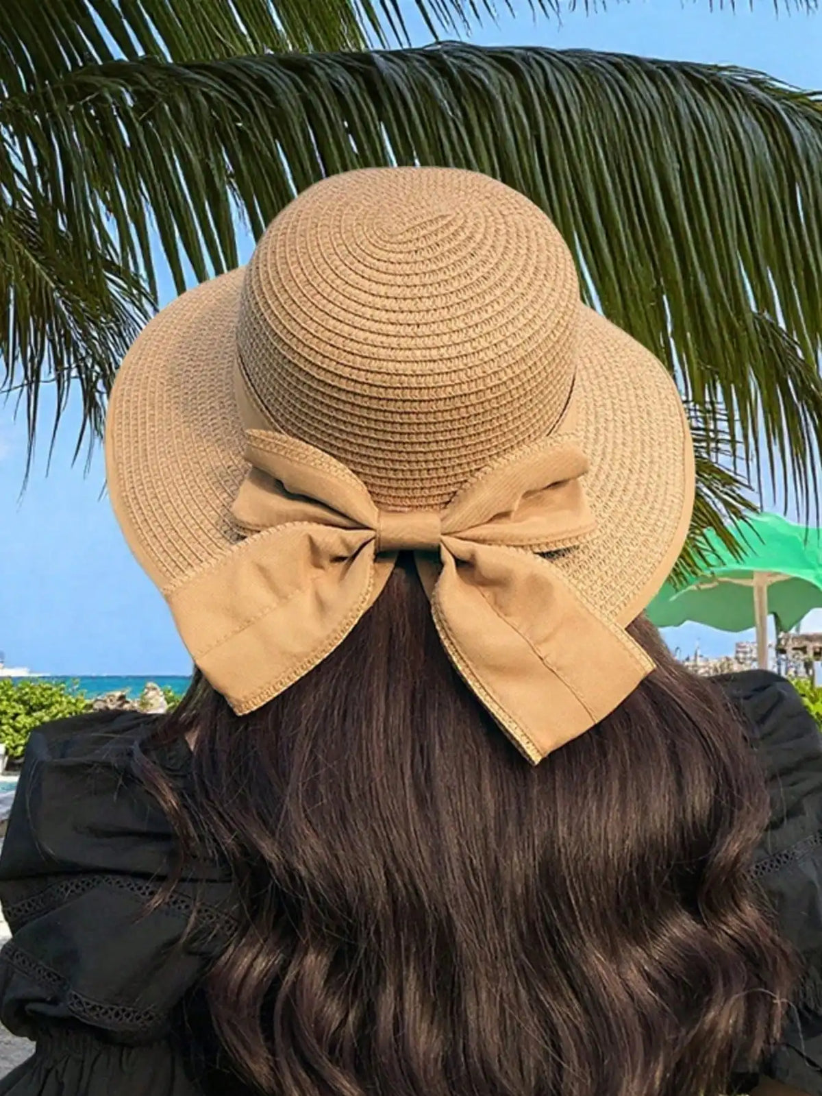 A woman wearing an elegant beige straw sun hat with a large bow on her dark hair against a tropical background.