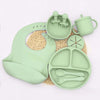 A light green silicone baby tableware set featuring a suction plate, bib, and various utensils on a woven mat.