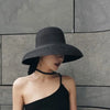 A woman wearing a wide brim straw sun hat and a black top against a gray stone wall.