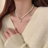 A close-up of a woman wearing an elegant necklace with a heart pendant and simulated pearls, paired with a cream-colored knit