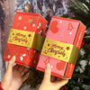 Rote Geschenkboxen mit Goldbuchstaben für ein Weihnachtsmann-Geldgeschenkset vor einem grünen Tannenbaum.