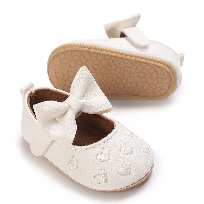 A pair of white soft non-slip baby first walker shoes with a bow detail and a tan sole.