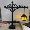A black wrought iron shelf lamp featuring an ornate cross design and a hanging lantern with a warm yellow glow.