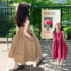 A young girl wearing a loose, sleeveless pink cotton sundress stands outdoors in a garden setting.