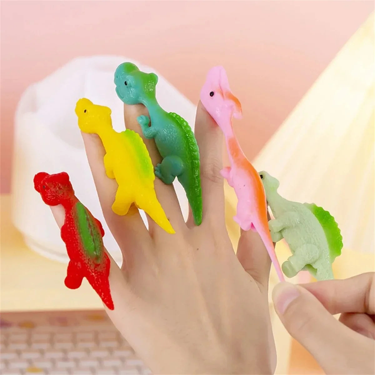 A hand holds several colorful dinosaur finger catapult toys, including green, yellow, pink, and red dinosaurs.