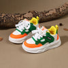 A pair of kids' breathable casual sneakers in green, orange, and yellow are displayed on a brown surface with a tree branch.