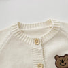 A cream colored knit cardigan with a round collar and buttons, featuring a cartoon bear design on the chest.