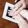 A hand is pressing a baby's handprint onto a white frame for an inkless keepsake kit.