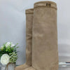 A pair of beige knee-high chunky heel boots with a metal buckle displayed next to white flowers.