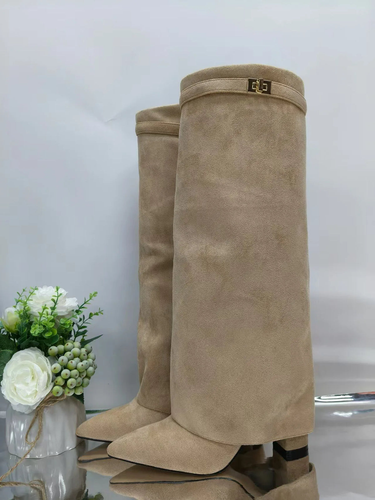 A pair of beige knee-high chunky heel boots with a metal buckle displayed next to white flowers.