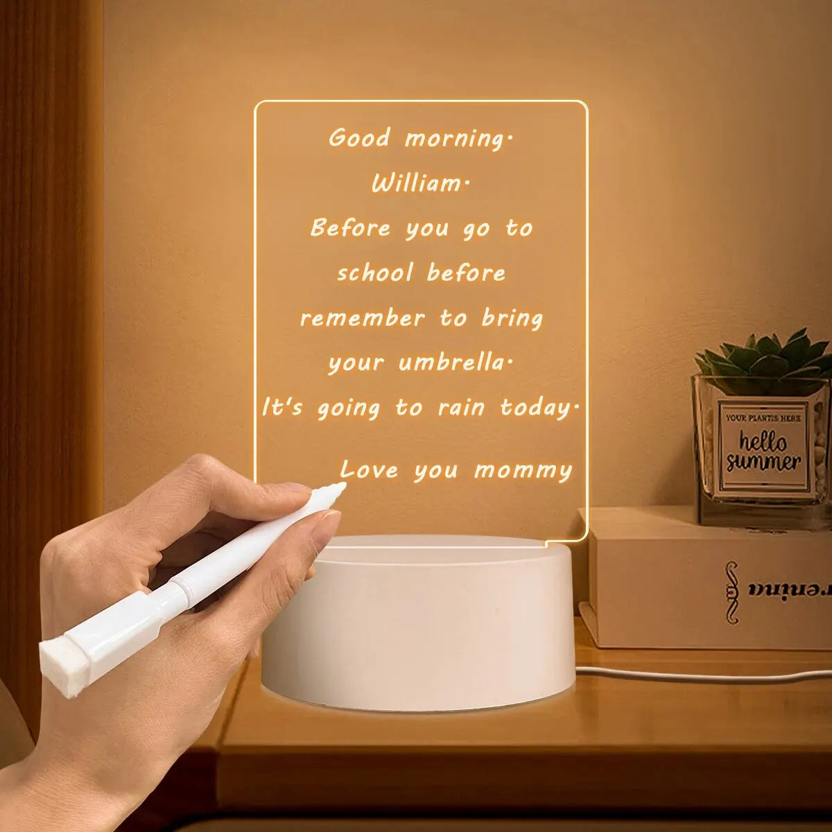 A hand writes a message on an acrylic message board lamp featuring a morning greeting and a reminder about an umbrella.