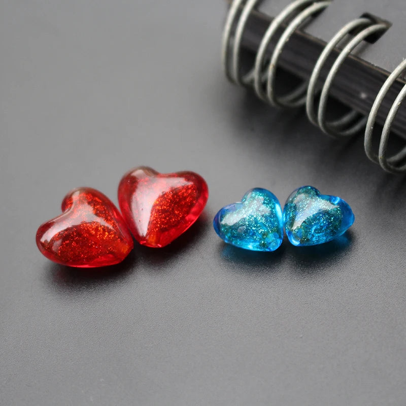 Deux perles de verre au chalumeau en forme de cœur, une rouge scintillante et une bleue, posées sur une surface sombre.