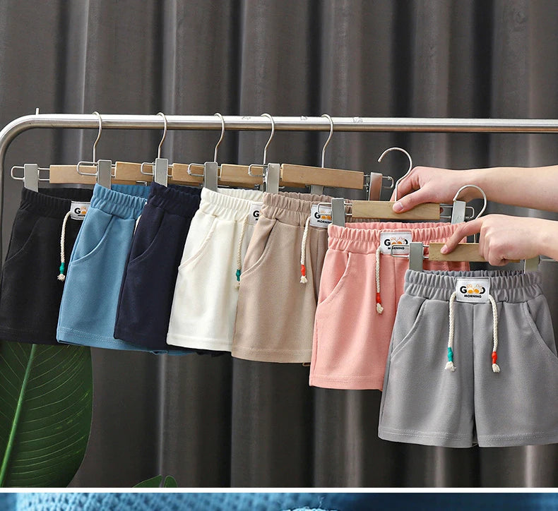 A hand is hanging various colored kids beach shorts on a clothes rack.