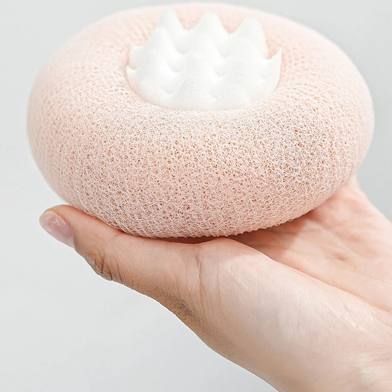 A hand holds a round, pink bath sponge with a white flower-like suction cup on top.