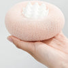 A hand holds a round, pink bath sponge with a white flower-like suction cup on top.