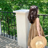 A girl wearing a loose, sleeveless, beige cotton sundress stands on a balcony with lush green foliage in the background.