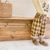 A child wearing beige checkered harem pants and a cream-colored top stands next to wooden steps.