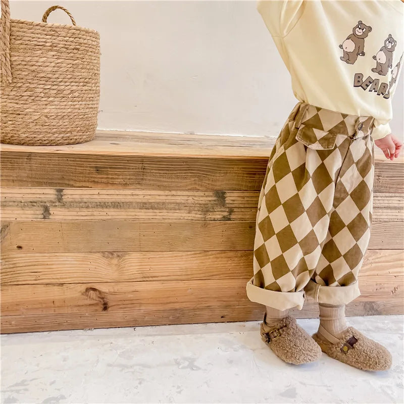 A child wearing beige checkered harem pants and a cream-colored top stands next to wooden steps.